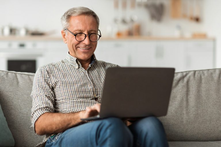 Happy Senior Man Using Laptop Browsing Internet Sitting At Home